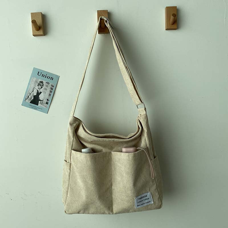 Beige corduroy crossbody bag with front pockets and adjustable strap, hanging on a wall hook.