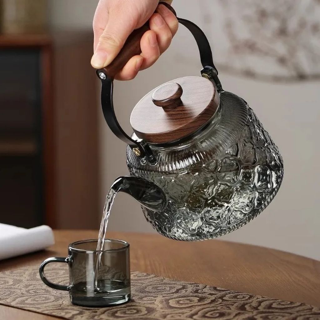 Hand pouring water from a grey patterned borosilicate glass teapot with a wooden lid and handle into a glass cup.