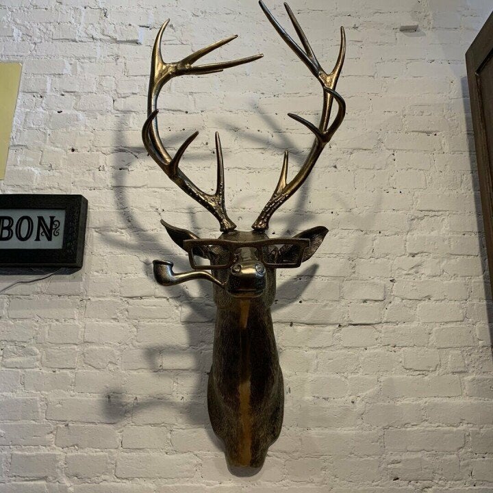 Bronze deer head wall decor wearing glasses and a pipe, mounted on a white brick wall.