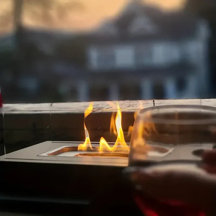 Fitlybuy smokeless black tabletop bio-ethanol fireplace with glass guards, glowing flame, and wine glass at sunset.
