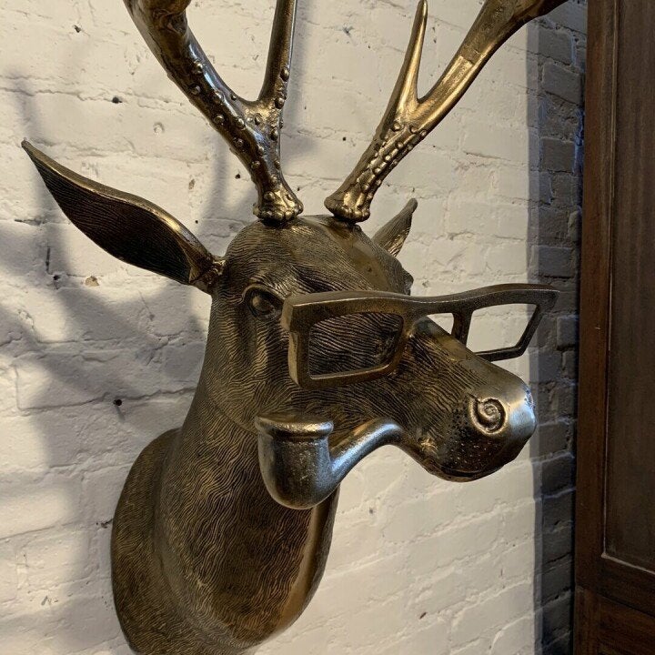 Gold-toned deer head wall decor wearing glasses and holding a pipe, mounted on a white brick wall.