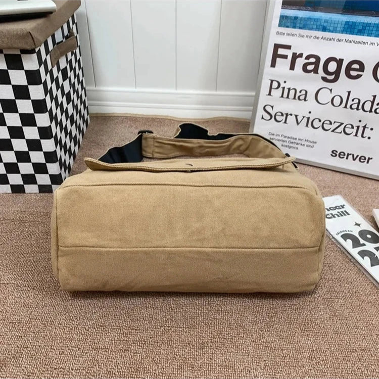 Tan Fitlybuy canvas messenger sling bag with black strap, positioned on a brown surface next to a checkered storage bin.