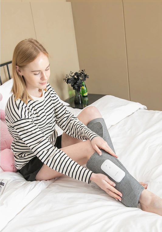 Woman sitting on a bed wearing a gray Fitlybuy air compression leg massager with a portable control unit.