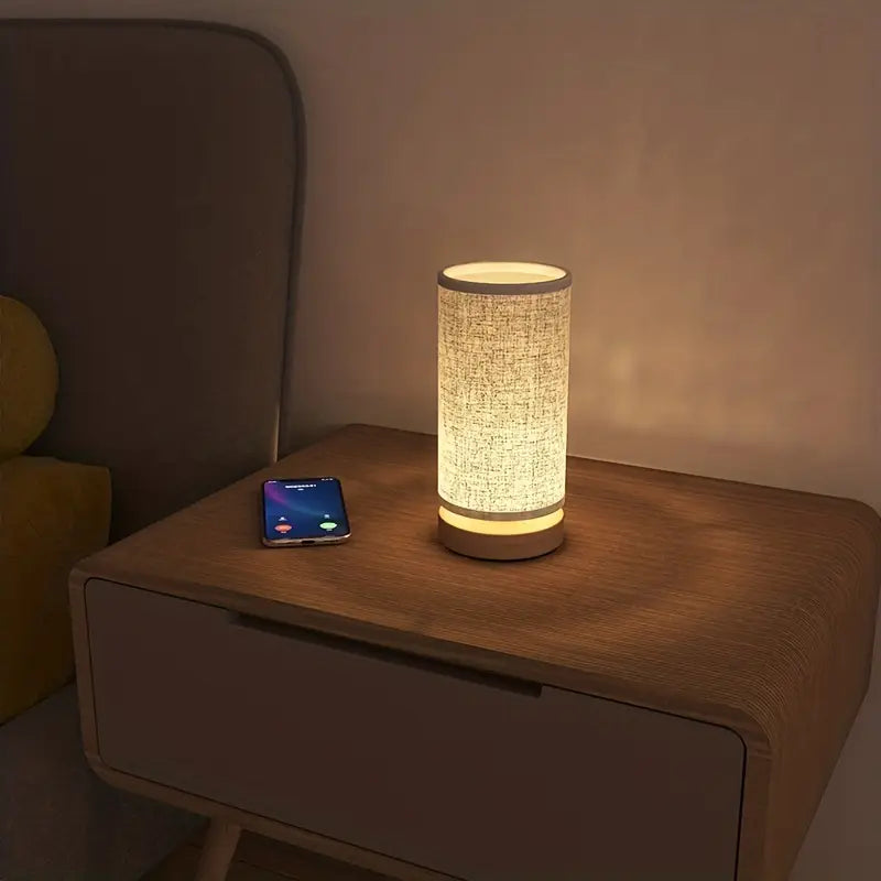 Fitlybuy minimalist cylindrical night lamp with fabric shade and wood base emitting warm light on a wooden nightstand.