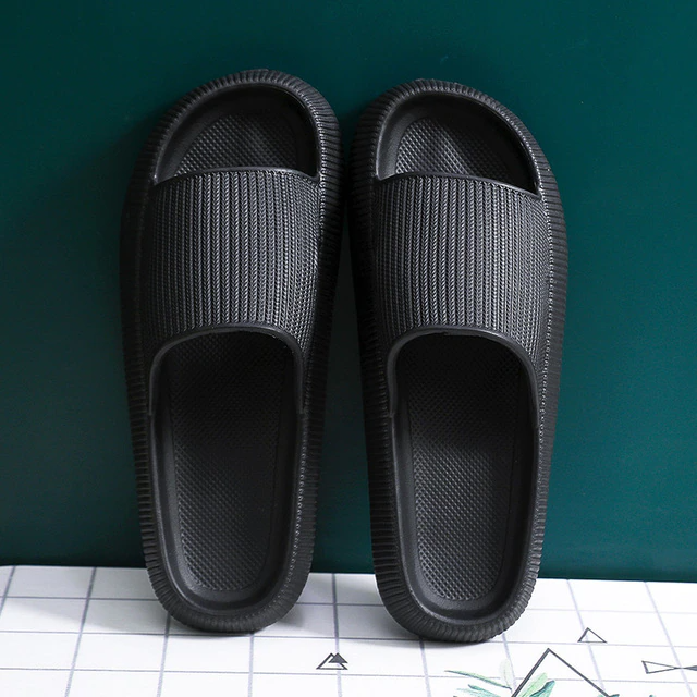 Pair of black Fitlybuy Cloud Slippers with textured memory foam design on a grid-patterned surface against green wall.