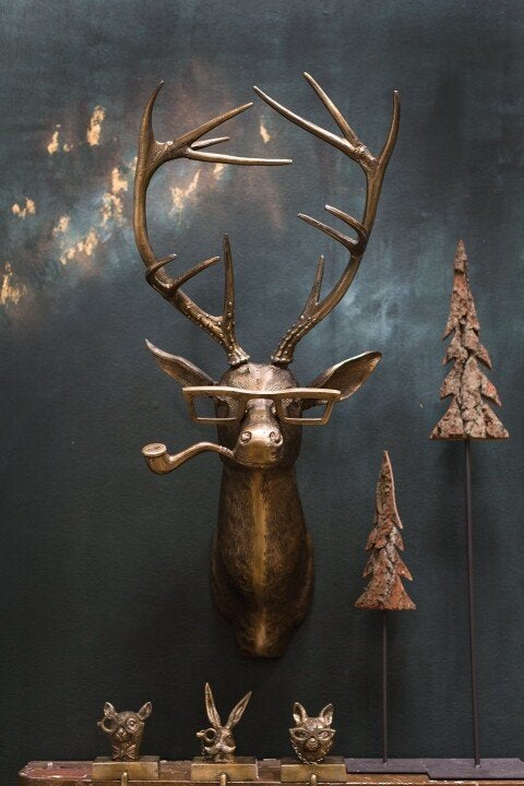 Bronze deer head wall decor with glasses and pipe, displayed on dark wall with miniature metal trees and animal figures.