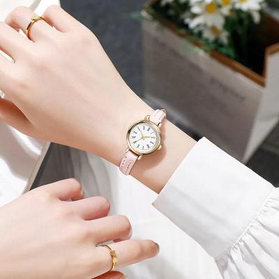 Fitlybuy Billie gold watch with round white face, Roman numerals, and thin pink leather strap on a woman’s wrist.