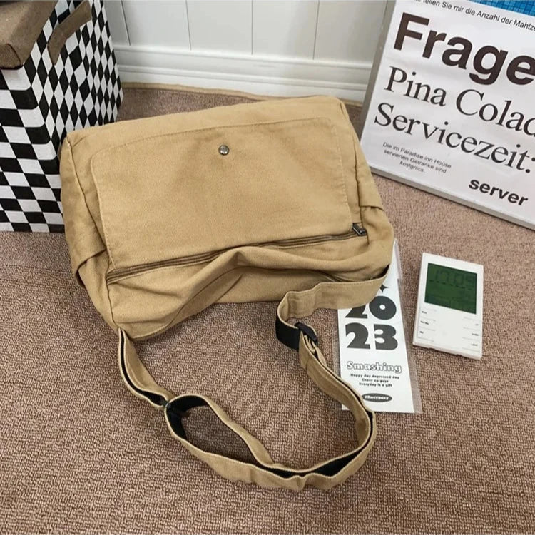 Tan canvas Fitlybuy CrossStream messenger sling bag with front snap pocket and adjustable strap on a textured floor.