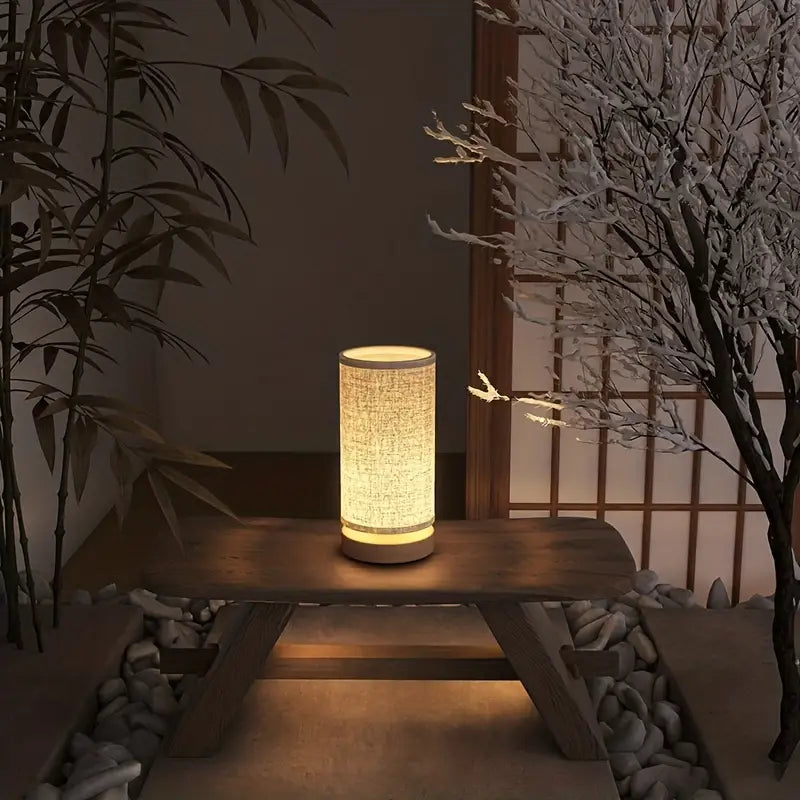 Fitlybuy SimpleGlow cylindrical wooden night lamp with a fabric shade emitting a warm, soft glow on a wooden bench.