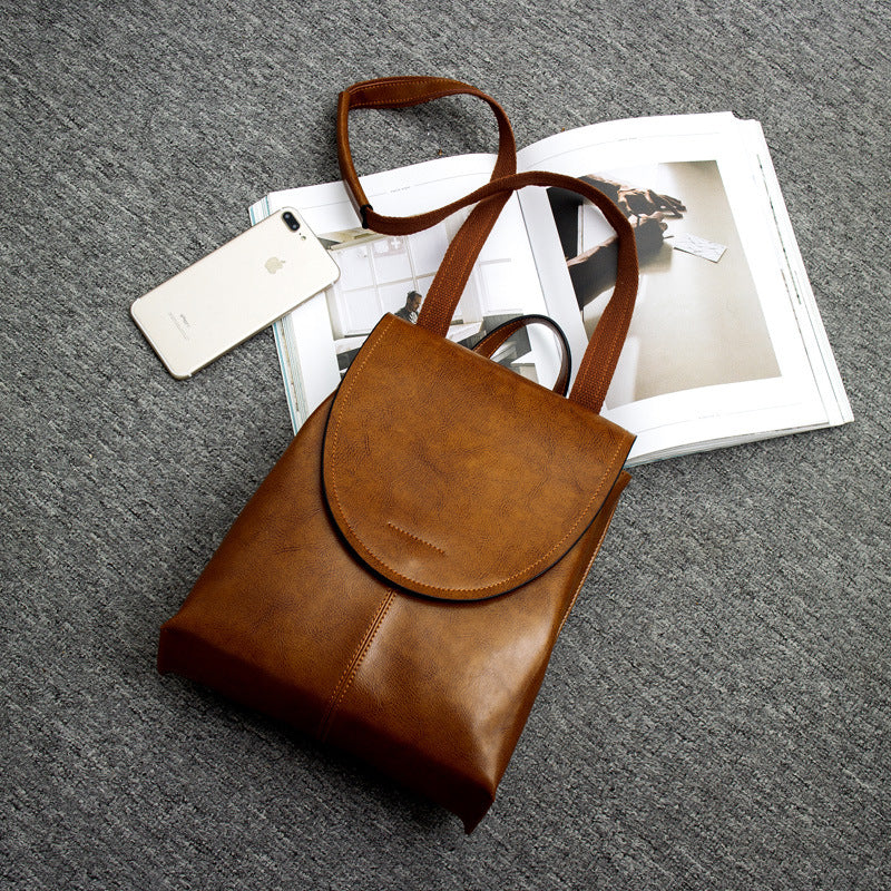 Fitlybuy brown leather backpack with a flap top and straps, displayed on a grey surface with a phone and open magazine.