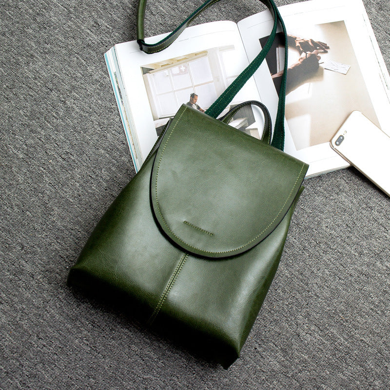 Olive green leather backpack with a flap closure, shown on a grey textured background with a magazine and smartphone.