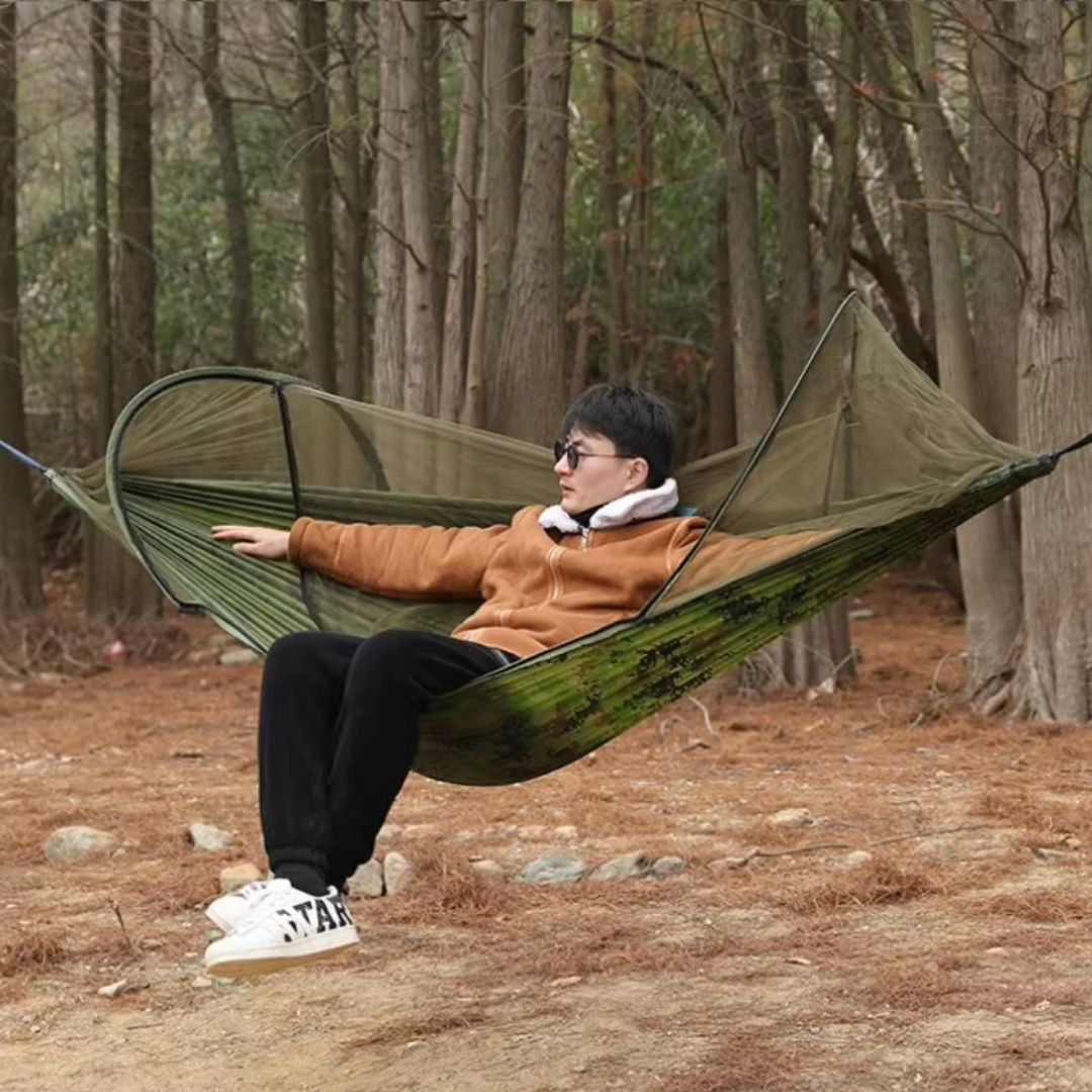Man in a green camouflage Fitlybuy hammock with an integrated BugShield mosquito net, set up in a wooded area.