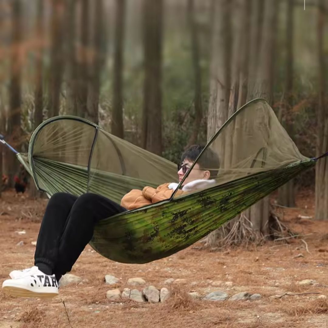 Person relaxing in a green camo Fitlybuy hammock with an integrated BugShield mosquito net in a wooded forest.