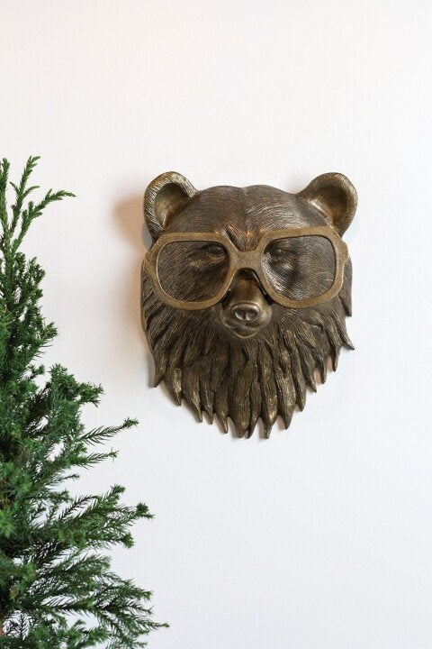 Bronze-colored bear head wall decor wearing glasses, mounted on a white wall next to a green pine branch.