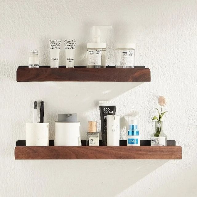 Two dark solid wood bathroom shelves mounted on a white wall, holding toiletries, toothbrushes, and a small flower.