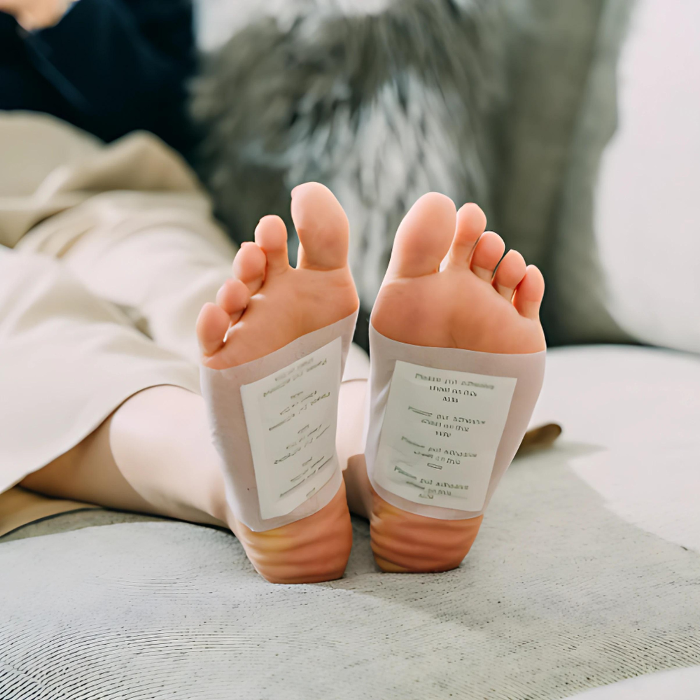 Feet with white Fitlybuy bamboo detox patches applied, resting on a sofa to show natural herbal infusion technology.
