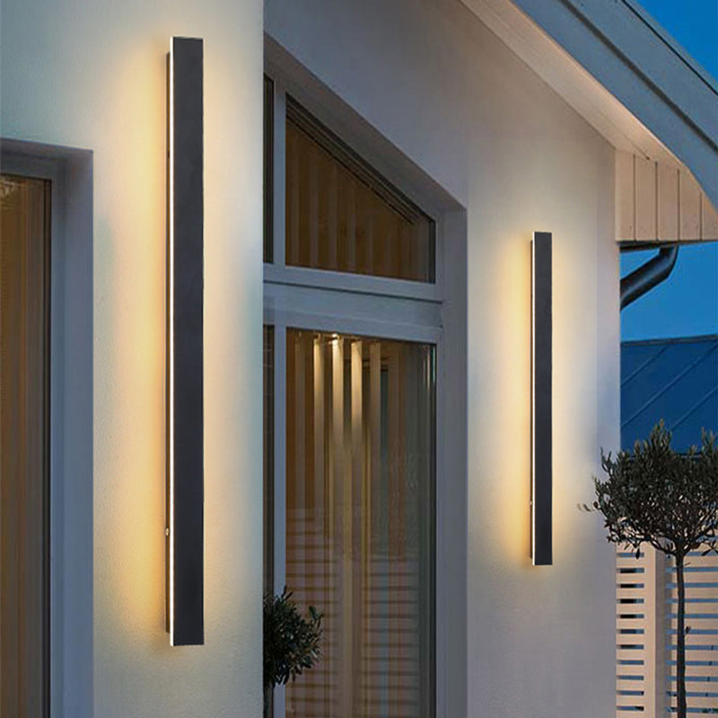 Two long, black, minimalist Fitlybuy LED wall lights casting a warm glow on a modern home exterior at dusk.