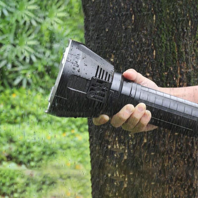 Hand holding a black Fitlybuy waterproof LED flashlight in heavy rain against a tree trunk.