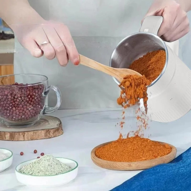 Fitlybuy electric grain mill pouring ground red spices onto a wooden plate, with beans in a glass mug nearby.