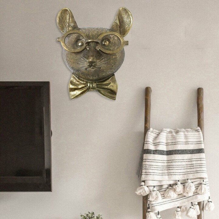Gold-toned, whimsical animal head wall decor wearing glasses and a bow tie, mounted on a neutral interior wall.
