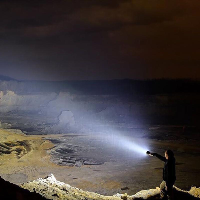 A person holds a powerful black LED flashlight, casting a long, bright beam over a dark, rocky quarry at night.
