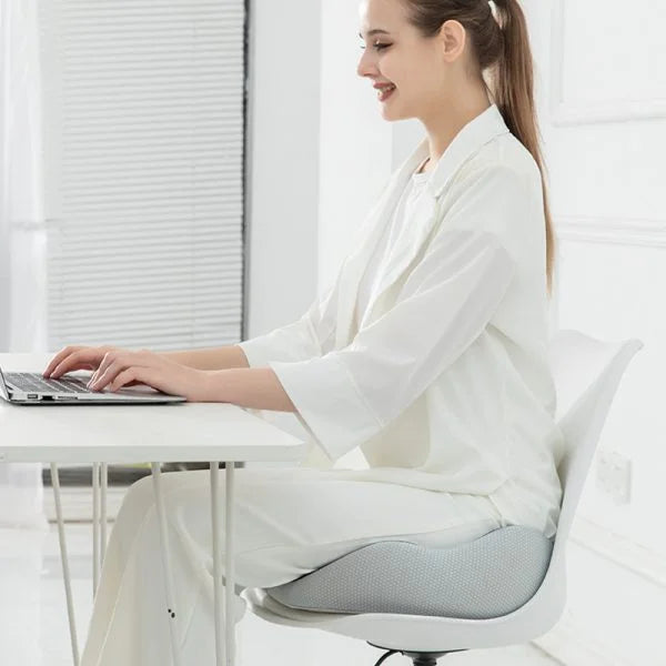 Woman sitting on a grey ergonomic Fitlybuy sciatica relief cushion on a white chair, typing on a laptop at a desk.
