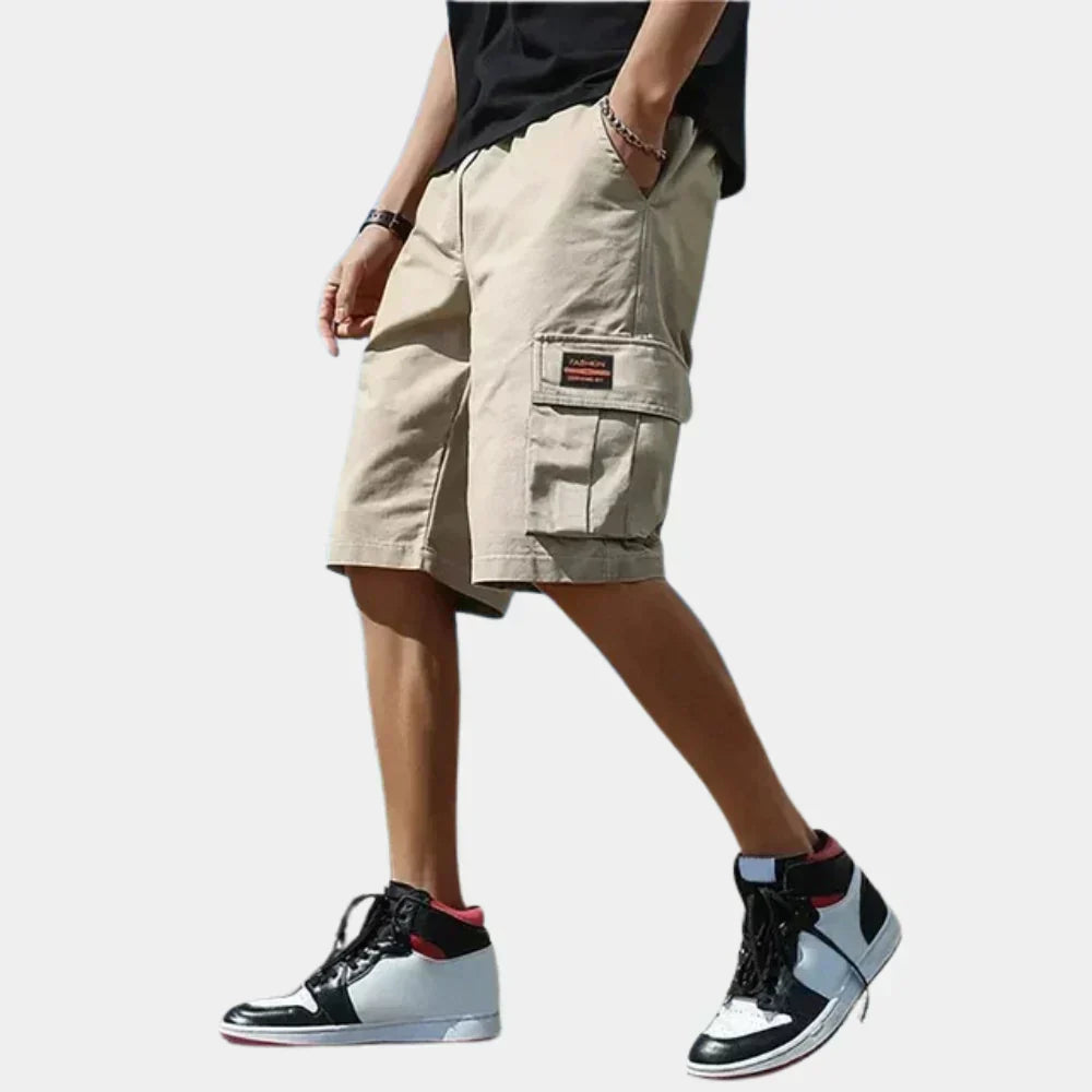 Fitlybuy men's loose-fit beige cotton cargo shorts with side pockets and a fashion patch, worn with high-top sneakers.