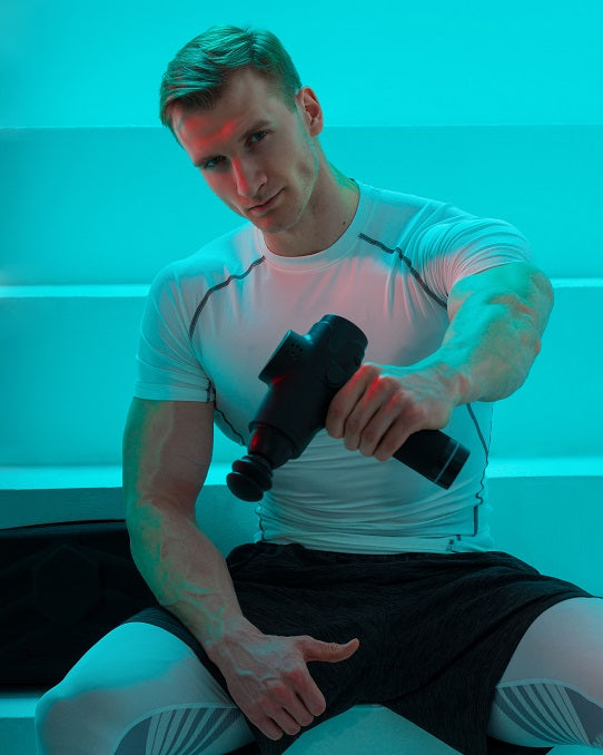 Fitlybuy ProMassage X1 black percussion massage gun held by an athletic man in a studio with teal lighting.