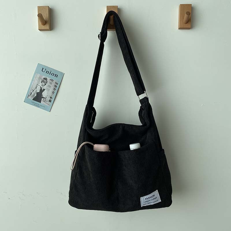 Black corduroy crossbody bag with adjustable strap and front pockets, hanging on a wall hook with a small art print.
