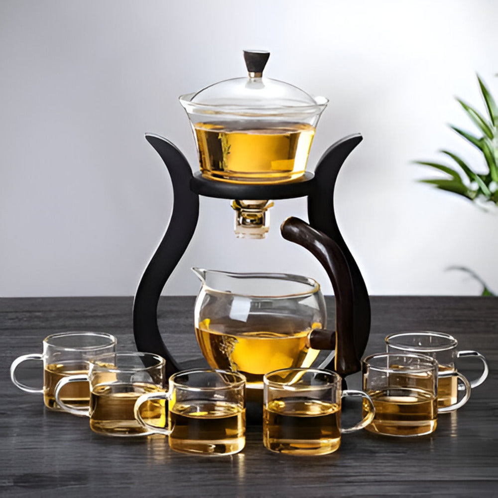Artisan magnetic glass teapot set with infuser on a black stand, surrounded by six glass cups filled with golden tea.