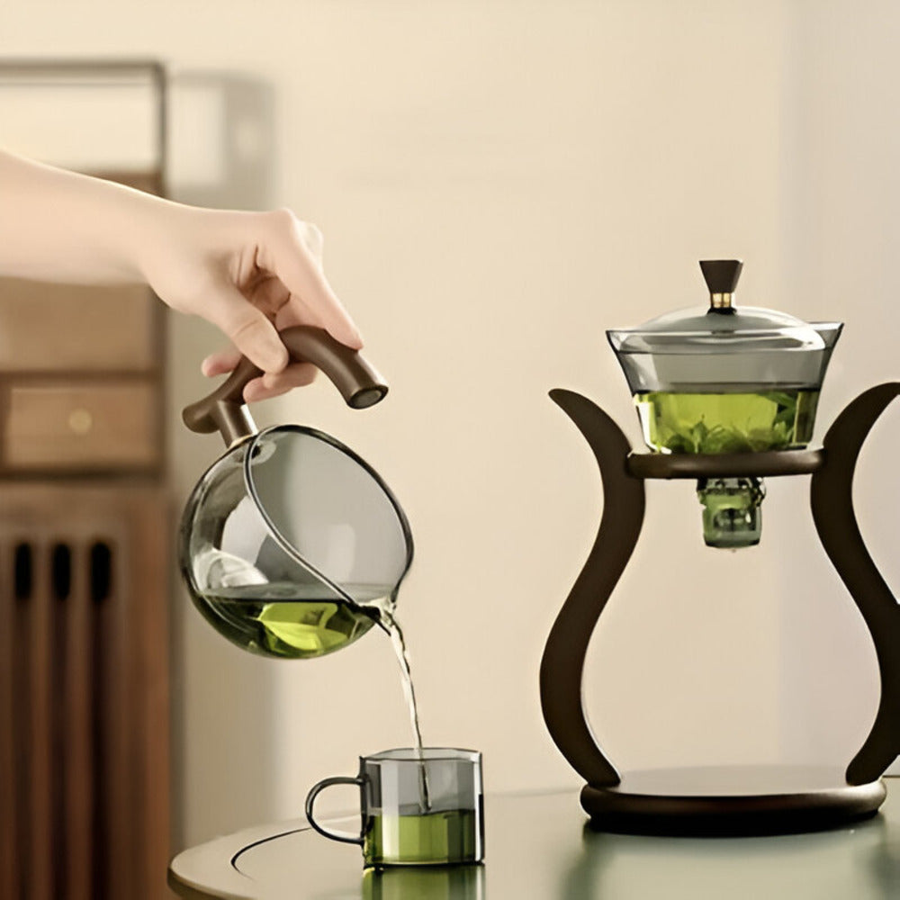 Hand pouring green tea from a Fitlybuy artisan magnetic glass teapot into a glass cup near a matching tea infuser.