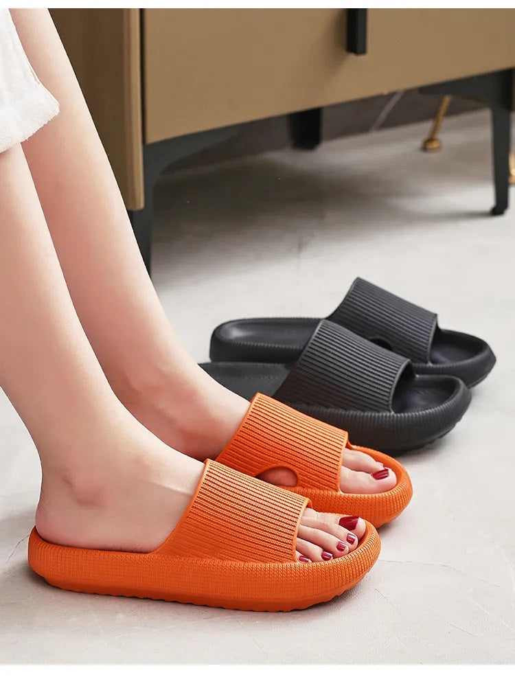 Fitlybuy cloud slippers in orange worn on feet, with a black pair in the background, featuring a thick, soft sole.