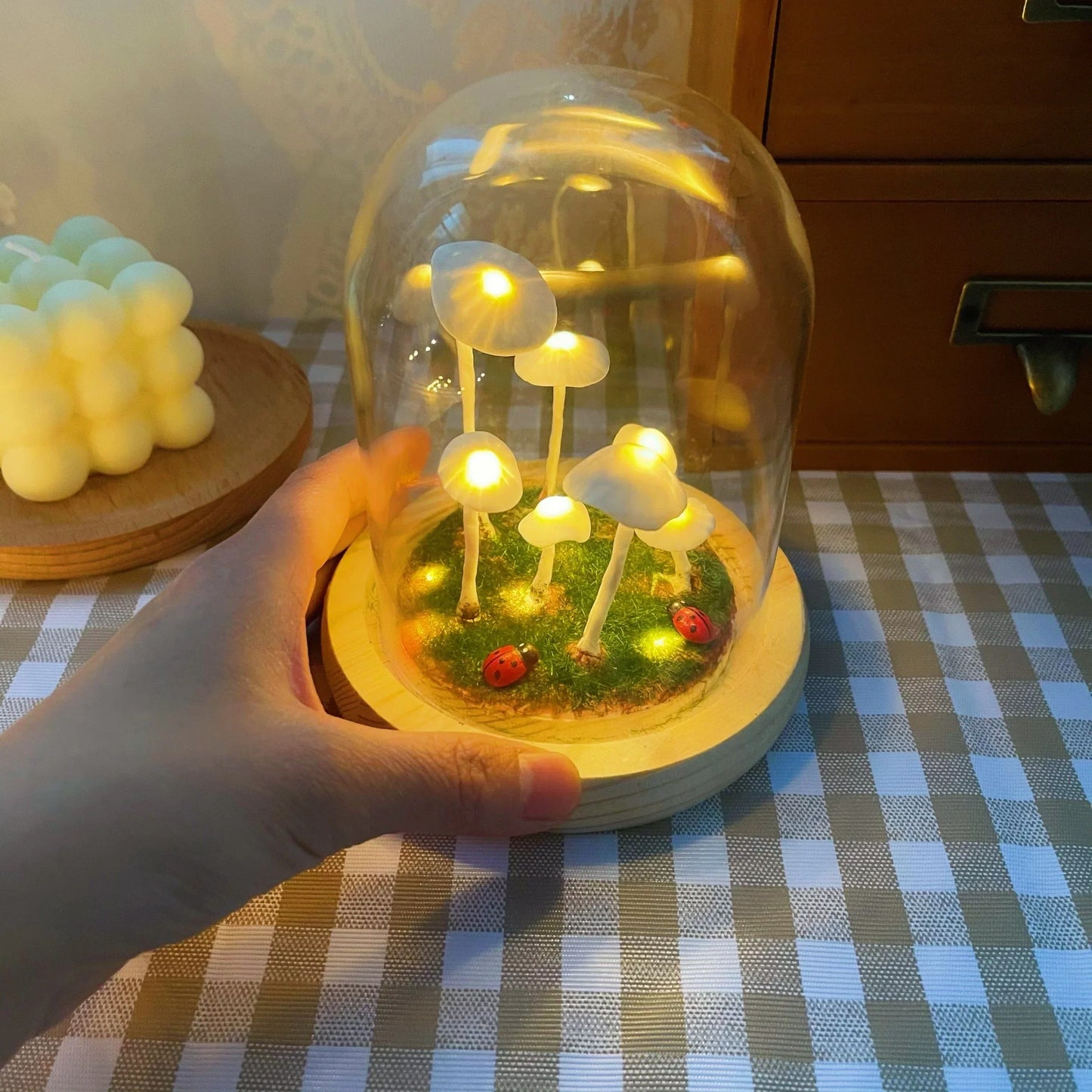 Hand holds a Fitlybuy glass lamp kit: white glowing mushrooms, ladybugs, and moss on a wood base on a checkered cloth.