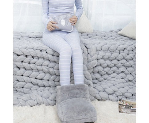 Person in striped loungewear using a soft gray Fitlybuy electric heated foot warmer while sitting on a chunky knit bed.