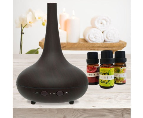 Dark wood-grain ultrasonic diffuser with Light and Mist buttons, beside three essential oil bottles on a spa table.
