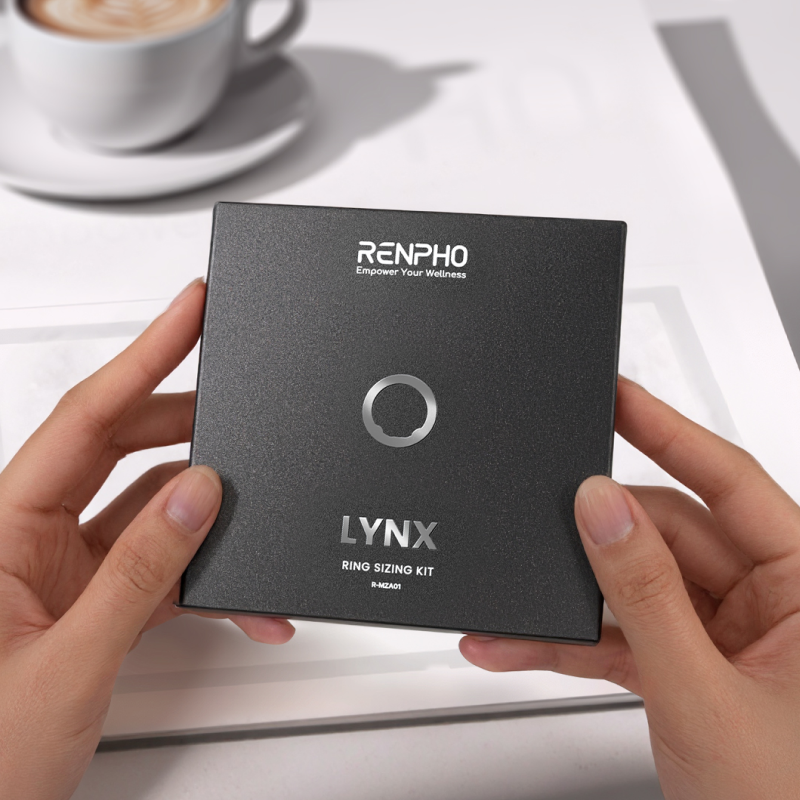 Hands holding a black Renpho LYNX smart ring sizing kit box with silver text and ring icon on a white desk with coffee.