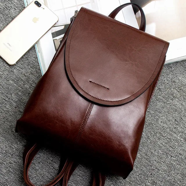 Fitlybuy brown leather backpack with a flap cover and top handle, set next to a smartphone on a gray surface.