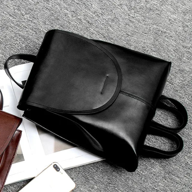 Fitlybuy black leather backpack with a flap cover and straps, displayed on a grey textured background with a smartphone.