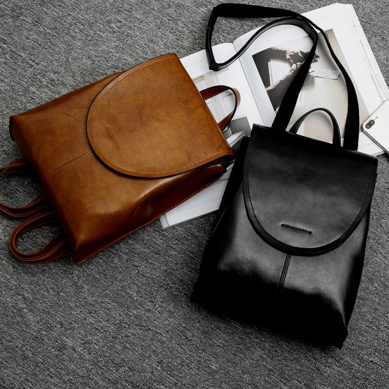 Brown and black Fitlybuy leather backpacks with fold-over flaps on a gray surface next to an open magazine.