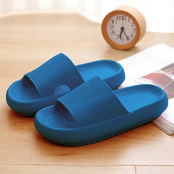 Blue Fitlybuy Cloud Slides with ComfortFoam cushioning on a wooden desk with a clock and notebook.