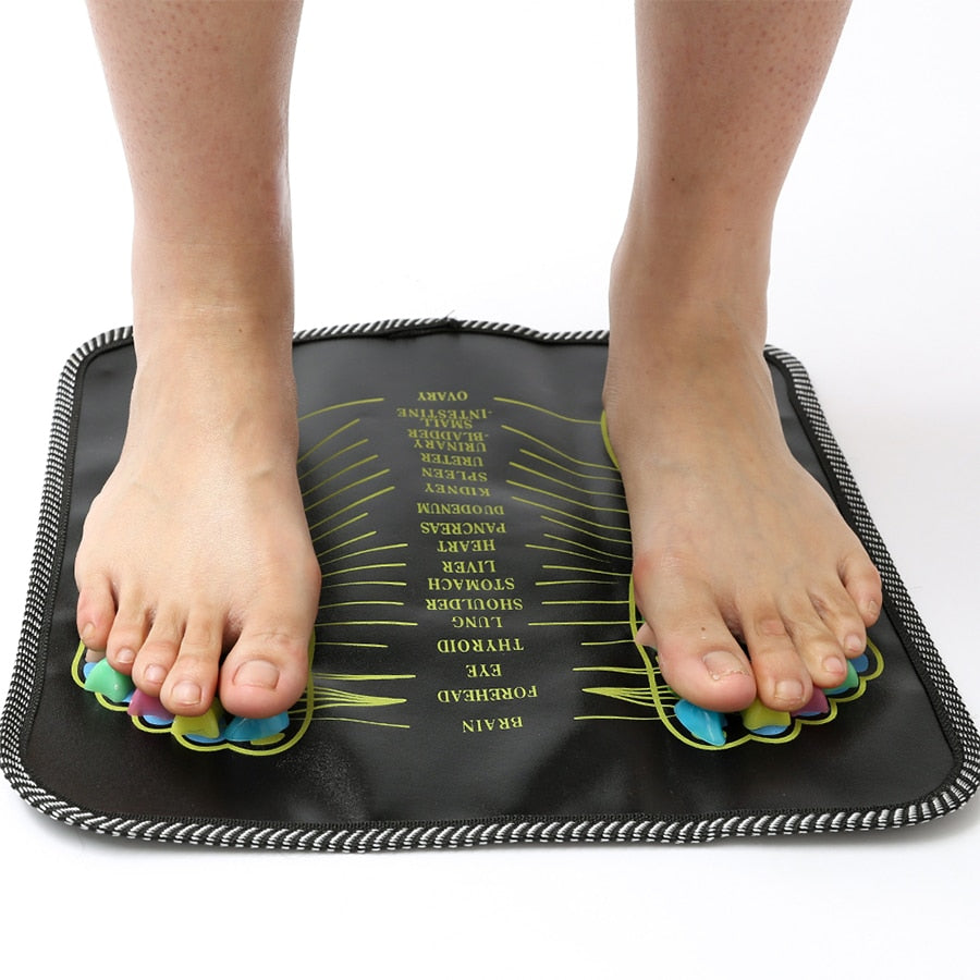 Feet standing on a black reflexology massage mat with colorful pressure point stones and printed body organ labels.