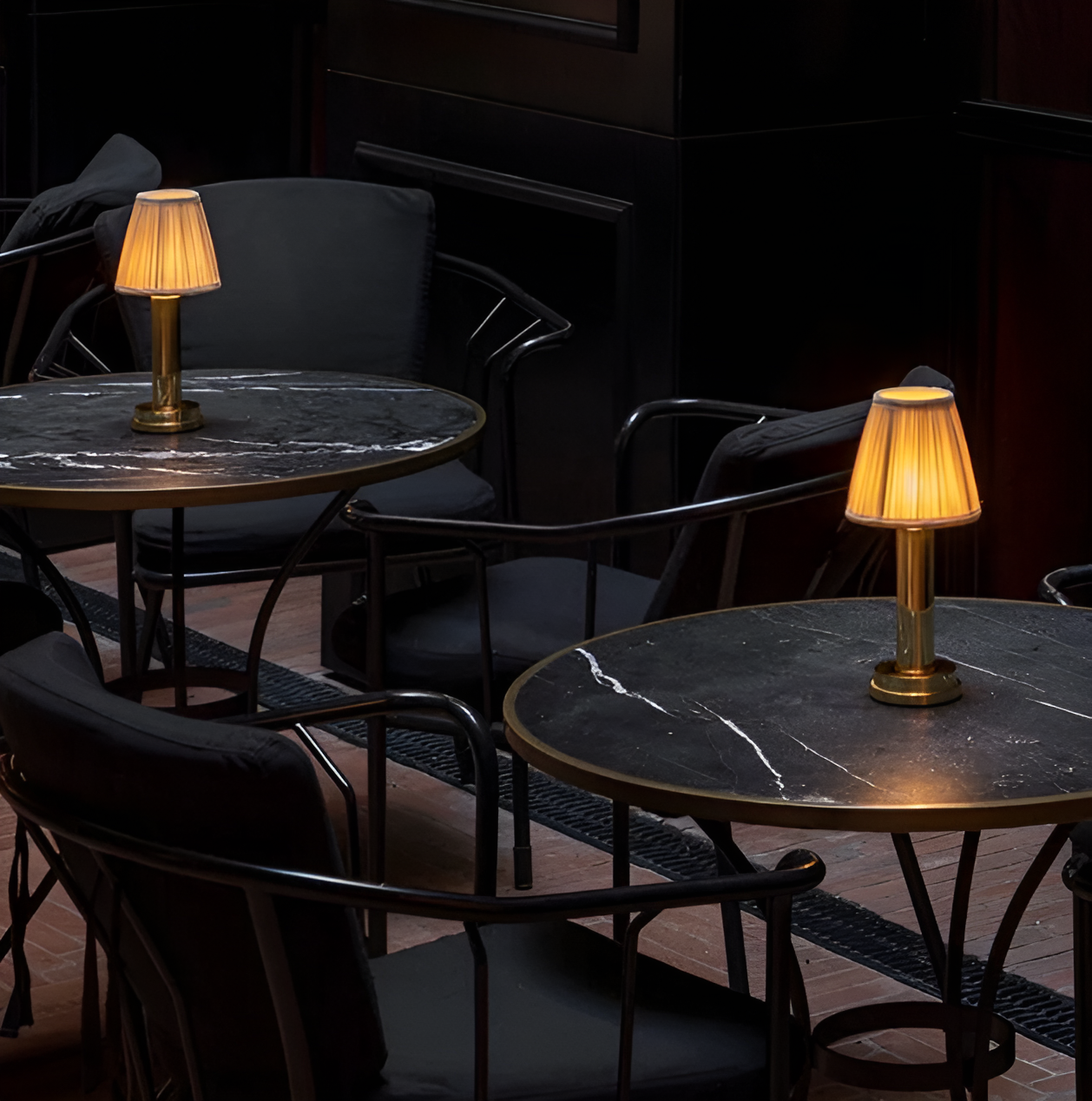 Two gold vintage flare table lamps with fabric shades, glowing on black marble bistro tables in a dark cafe.