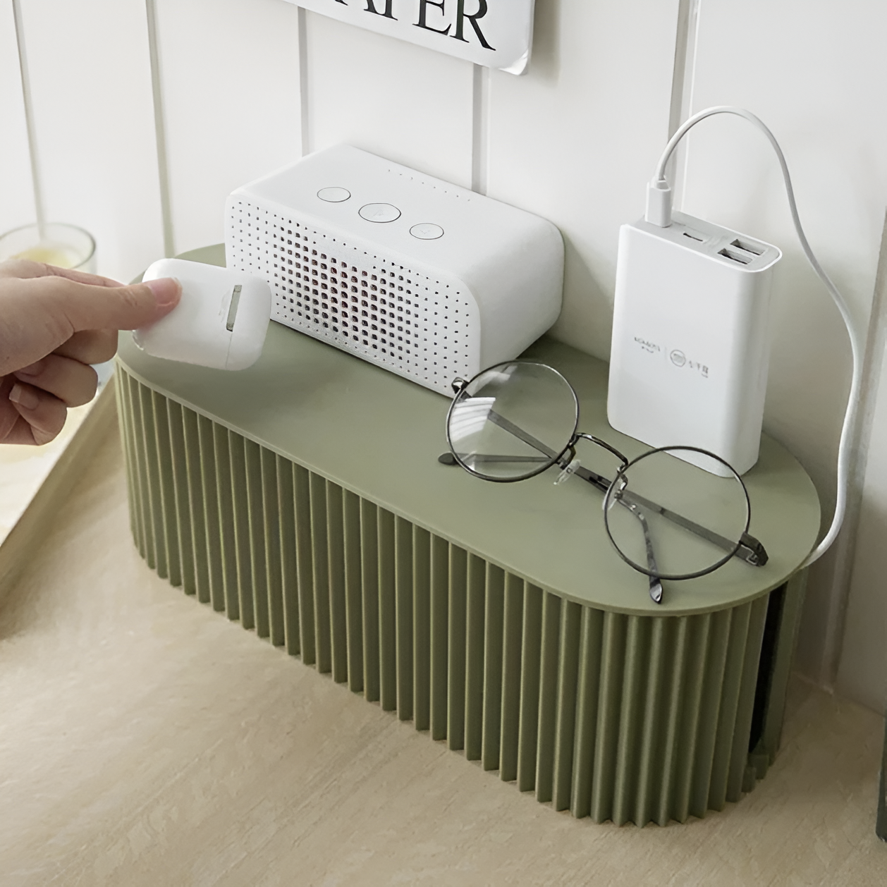 Sage green oval cable organizer box with a ribbed texture, acting as a desktop stand for electronics and accessories.