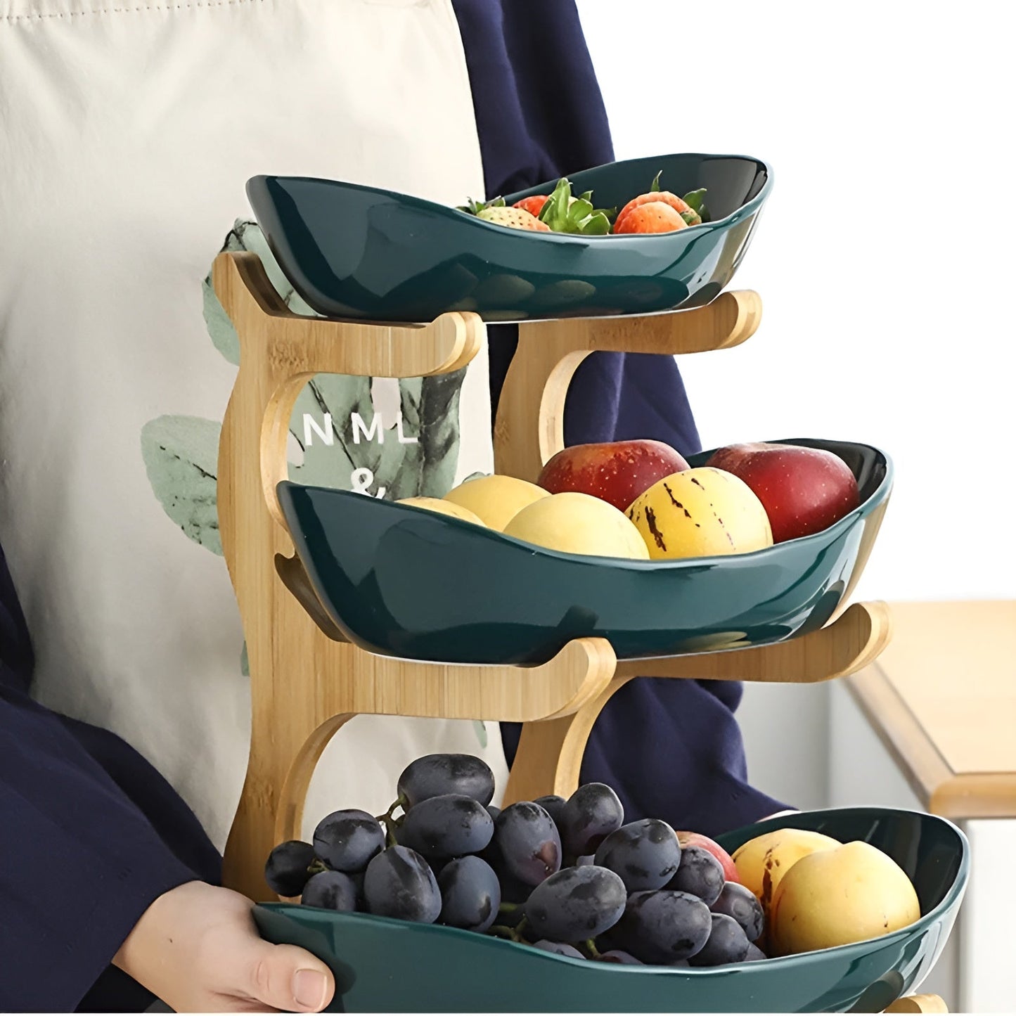 Person holding a Fitlybuy 3-tier wooden stand with dark green ceramic bowls filled with assorted fresh fruit.