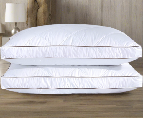 Two white, plush king-size pillows with diamond-stitched patterns, stacked on a wooden floor in a bedroom setting.