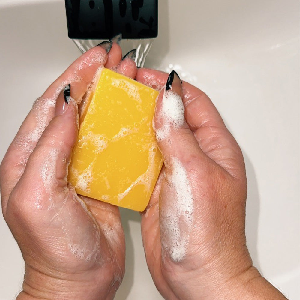 Hands holding a bright yellow Fitlybuy turmeric and kojic acid brightening soap bar covered in white lather under water.