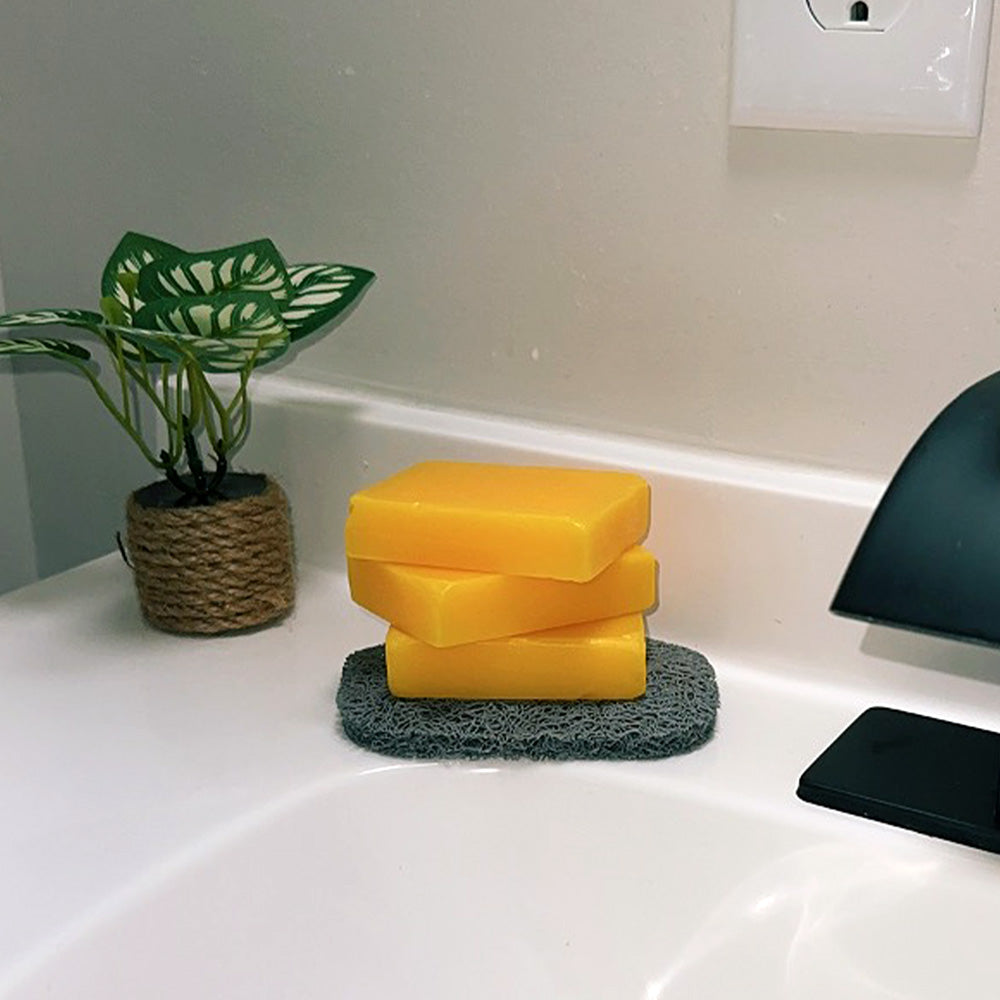 Fitlybuy yellow turmeric and kojic acid soap bars stacked on a grey soap dish next to a small plant on a vanity.