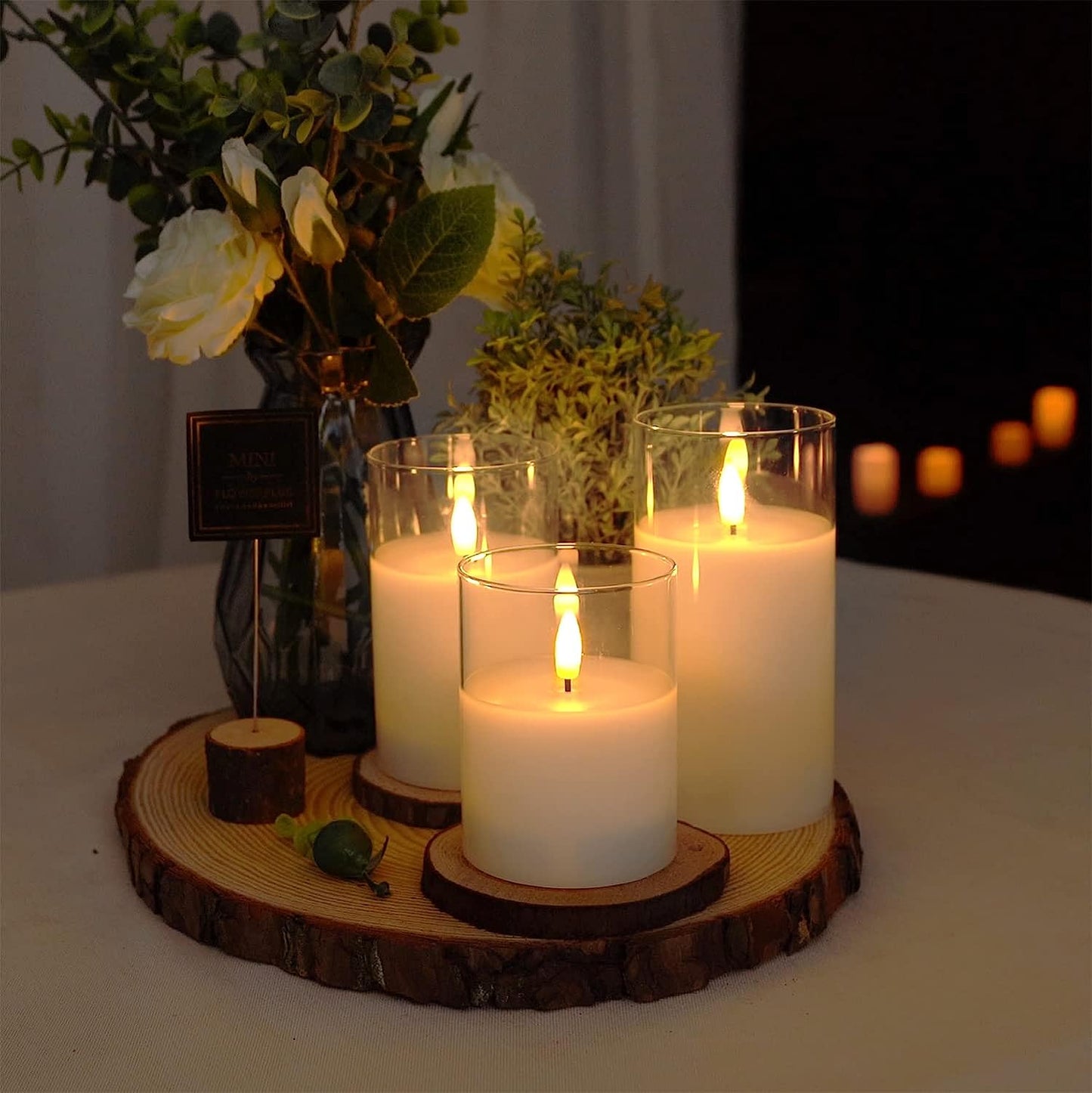 Fitlybuy AuraFlame LED candles in glass holders on a wooden base with faux flowers, creating a warm, realistic ambiance.