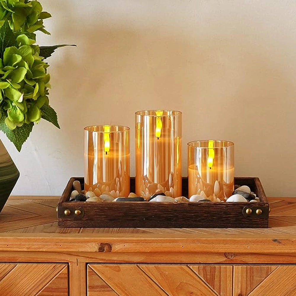 Fitlybuy AuraFlame LED candles in gold glass sleeves, set on a decorative wooden tray with stones on a wooden console.