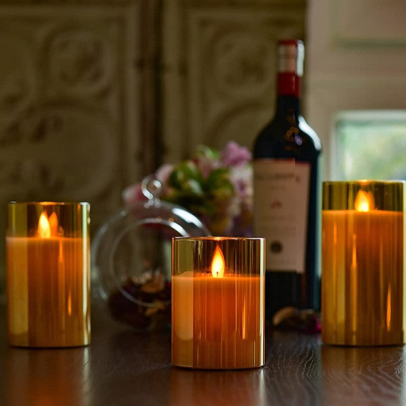 Three Fitlybuy AuraFlame LED candles in gold glass sleeves with realistic flames on a table with wine and decor.